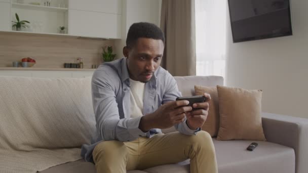 Joven adulto negro macho está jugando un juego en su teléfono inteligente y falla una ronda con una reacción brillante a ella — Vídeo de stock