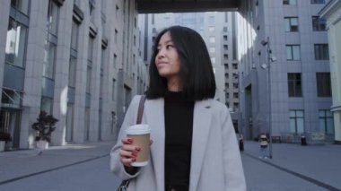 Genç Asyalı iş kadını elinde kahve fincanıyla ağır çekimde ofisten çıkıyor.