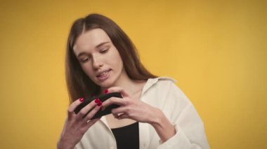 Genç beyaz kadın akıllı telefonuyla online video oyunu oynuyor. Parlak sarı arka plan.