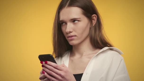 Νεαρή λευκή γυναίκα σκέφτεται μια απάντηση στο έξυπνο τηλέφωνό της σε ένα φωτεινό κίτρινο φόντο — Αρχείο Βίντεο
