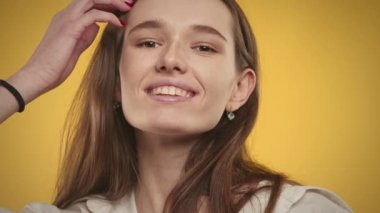 20 'li yaşlarda beyaz bir kadının parlak sarı bir arka planda kameraya gülümseyen portresi.