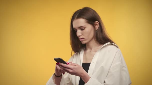 Νεαρή λευκή γυναίκα εκπλήσσεται από μια συμφωνία στο smartphone της σε ένα φωτεινό κίτρινο φόντο — Αρχείο Βίντεο
