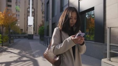 Genç Asyalı kadın şehirde yürürken akıllı telefonuyla yazı yazıyor.