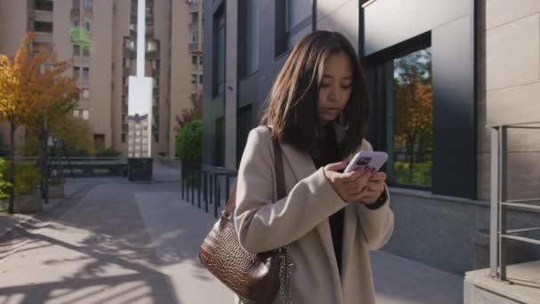 Молодая взрослая азиатка печатает на смартфоне во время прогулки по городу — стоковое видео