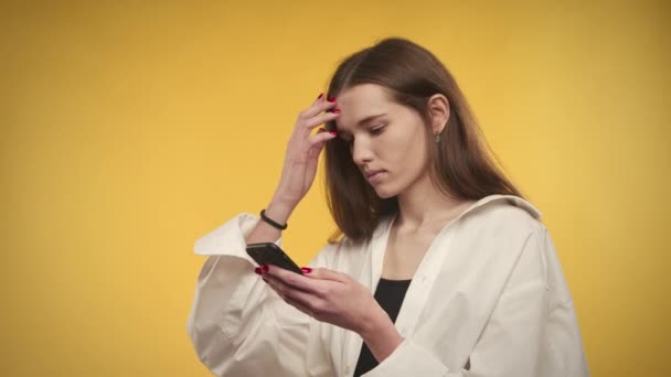 Joven mujer caucásica adulto está navegando en su teléfono inteligente sobre un fondo amarillo brillante en el estudio — Vídeos de Stock