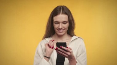 Genç beyaz kadın, parlak sarı bir arka planda akıllı telefonuna bakarken meraklı.