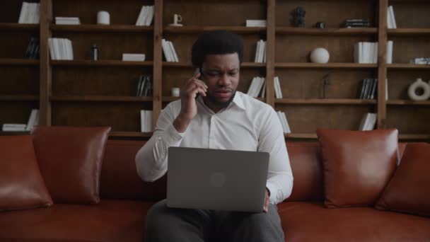 若い大人のビジネスマンは彼の前でノートパソコンと彼の携帯電話で否定的な会話をしています — ストック動画