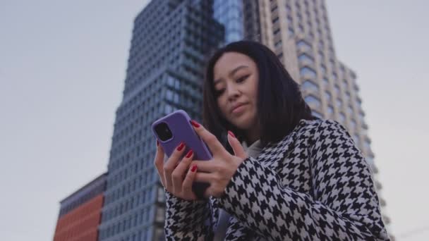 Junge erwachsene asiatische Frau surft auf ihrem Smartphone unter einem Hochhaus — Stockvideo