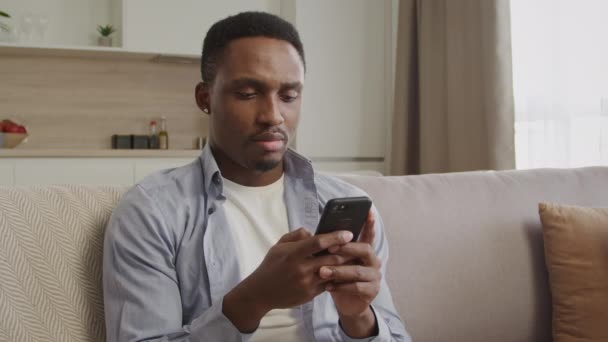 Portret van een Afrikaan die denkt en typt op een smartphone in zijn appartement. — Stockvideo