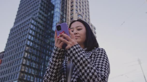 Mujer adulta joven leyendo en su teléfono inteligente bajo un edificio de gran altura en el centro de la ciudad — Vídeos de Stock