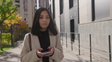 Genç Asyalı iş kadını akıllı telefonuyla yazı yazıyor ve şehirde yürürken gözlerini kaçırıyor.