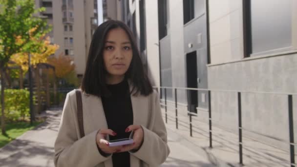 Junge erwachsene asiatische Geschäftsfrau tippt auf ihrem Smartphone und schaut weg, während sie in der Stadt spaziert — Stockvideo