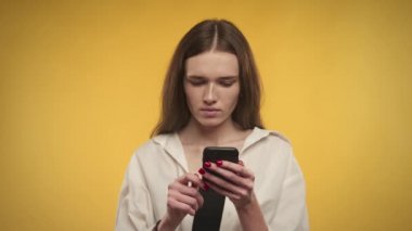 Genç beyaz kadın, parlak sarı bir arka planda akıllı telefonundan harika haberler alıyor.