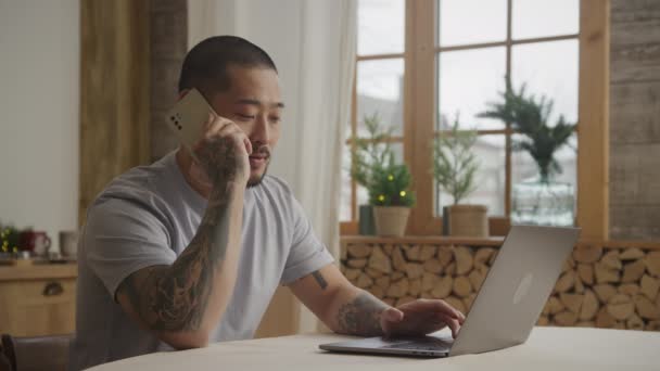 Young adult asian enterpreneur performing a sale while talking on his phone in front of his laptop — Stockvideo