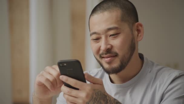 Junge erwachsene asiatische Mann surft auf seinem Smartphone mit nicht schlecht Gesichtsausdruck drinnen — Stockvideo