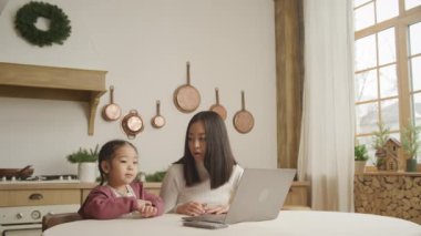 Evde dizüstü bilgisayarın önünde konuşan genç bir anne ve kız.