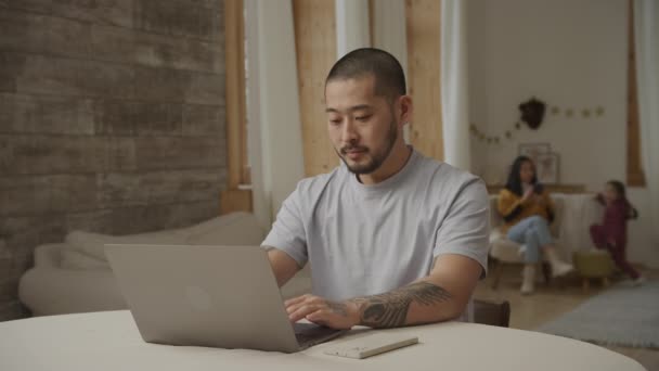 Ein glücklicher Vater arbeitet an einem Laptop, während seine Familie drinnen im Hintergrund spielt — Stockvideo