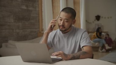 Genç baba, dizüstü bilgisayarının önünde ailesi arkasındayken telefona cevap veriyor.
