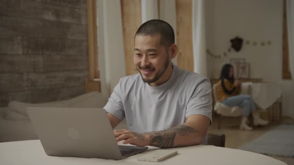 Lycklig far arbetar med ett leende på en bärbar dator med sin familj i bakgrunden — Stockvideo