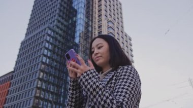 Şehir merkezindeki akıllı telefonuna bakan genç bir kadın. Dışarıdaki gökdelenin altında.