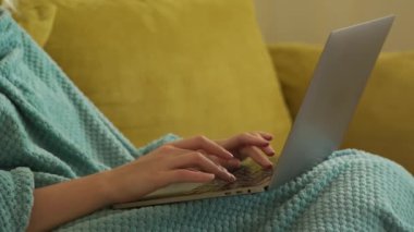 Close up of a caucasian woman hands typing on a laptop computer under a blue blanket on a yellow sofa