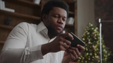 Yakın plan portresi Afro-Amerikan erkek çevrimiçi akıllı telefonla oynuyor. Noel ağacı arka planı yavaş çekimde kapalı alanda.