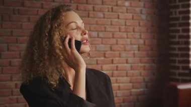 Genç, kıvırcık, sarışın, 30 'lu yaşlardaki iş kadını arka planda kırmızı tuğla bir duvarla çatı katında telefonla konuşuyor.