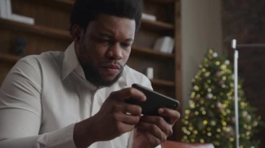 Close-up portrait African-American man gaming on smart phone online with Christmas Tree background in slow motion