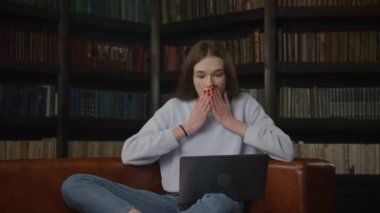 Young adult caucasian woman recieving good news on her laptop computer in front of bookshelf indoors