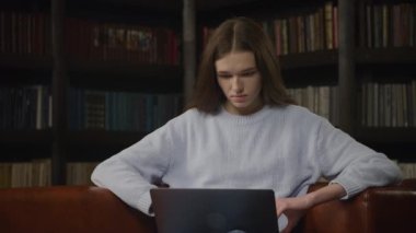Young adult caucasian woman student working on a laptop computer indoors in a bookshelf room in slow motion