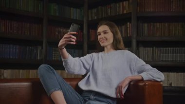 Young adult caucasian woman student taking selfie on her smart phone indoors in slow motion with a bookshelf behind