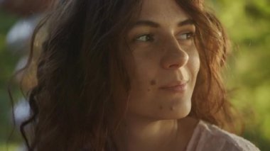 Close up portrait of a young adult curly caucasian woman with freckles looking at sunlight during sunset shot handheld in slow motion
