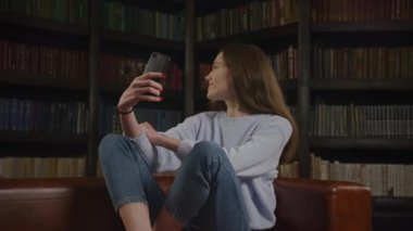 Young adult caucasian woman student taking selfie on her smart phone indoors in slow motion with a bookshelf behind