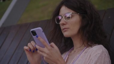 Handheld approaching shot of a curly caucasian woman browsing on her purple smart phone on a bench in the park in slow motion 90fps