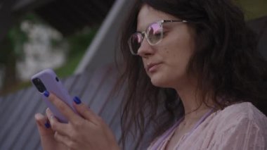Handheld approaching shot of a curly caucasian woman browsing on her purple smart phone on a bench in the park in slow motion 90fps