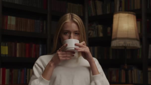 一名穿着白色毛衣和一本书在屋里喝茶或喝咖啡的年轻成年女子的近身镜头 — 图库视频影像