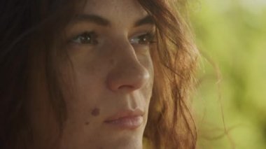 Handheld close up catching sunbeam on curly caucasian woman face with freckles and birthmark outside in slow motion