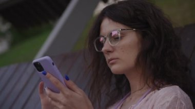 Handheld approaching shot of a curly caucasian woman browsing on her purple smart phone on a bench in the park in slow motion 90fps