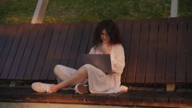 Handheld shot of a curly caucasian woman woking on her laptop computer on a park bench on sunset in slow motion
