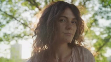 Pivoting around close up of a young adult curly caucasian woman looking at camera with lens flare from setting sun