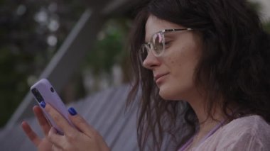 Handheld approaching shot of a curly caucasian woman browsing on her purple smart phone on a bench in the park in slow motion 90fps