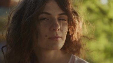 Pivoting around close up portrait of a young adult curly caucasian woman with freckles looking at camera in sunset sunlight in slow motion