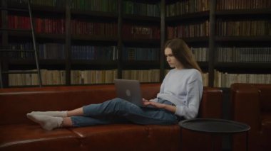 Dizüstü bilgisayarı üzerinde ağır çekimde çalışırken kanepede dinlenen beyaz bir kadın.