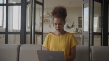 Dizüstü bilgisayarda çalışan girişimci. Bilgisayar kullanan kadın. Genç, Afro-Amerikan çalışanı mutlu bir şekilde dairede çekilen ağır çekimde işine odaklanmış.