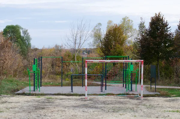Yeşil ve siyah metal ekipmanlı bir açık hava spor sahası ve futbol sahasının bir kısmı, parkta sonbahar ağaçlarıyla dolu.. 