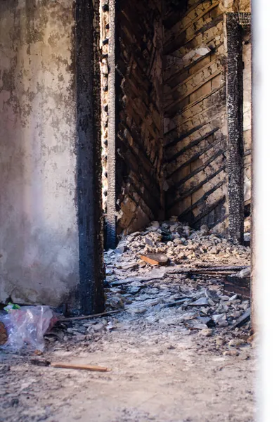  Yanmış evin kalıntıları. Hasarlı duvarlar, kayalar ve yerdeki çöpler. Güneş ışığı duvardaki delikten geliyor.. 