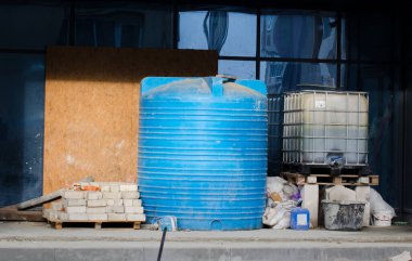 İnşaat alanında. Büyük mavi plastik depolama tankı, gri tuğla, kontrplak ve fütüristik cam pencere arka planda.