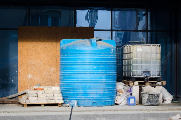 İnşaat alanında. Büyük mavi plastik depolama tankı, gri tuğla, kontrplak ve fütüristik cam pencere arka planda.