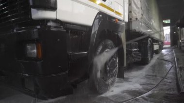 Kamyonun ön kısmı güçlü bir su püskürtüsüyle kenardan sulanır. Kamyonun farları ve bir tarafı görünür.