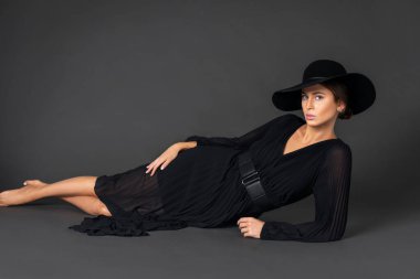 Portrait of an elegant young white woman with wavy hair and beautiful makeup laying by herself on the floor inside a studio with a grey background wearing a black dress and hat.